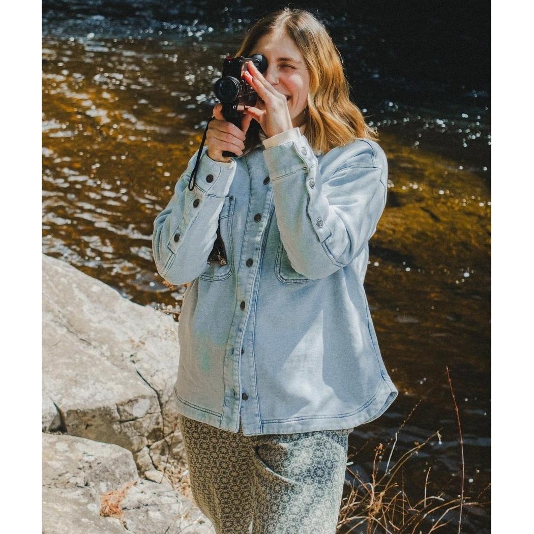 A woman wearing the Jetty Women's Eden Knit Shirt in Denim holds a camera by a river.