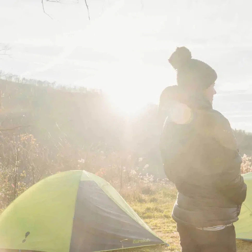 A person in a beanie stands near the Johnson Outdoors Eureka! Suma 2 tent in a sunlit, grassy outdoor setting.