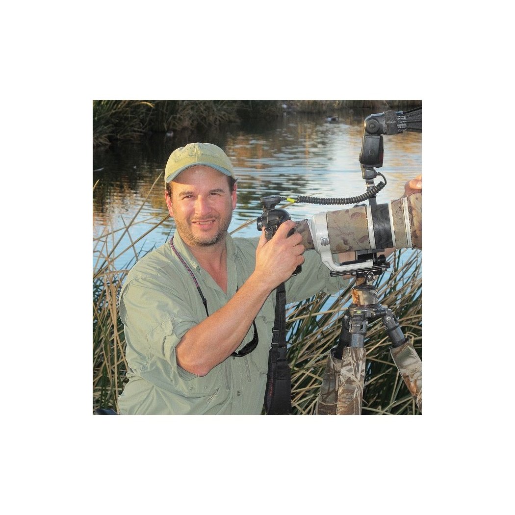 A man outdoors by a lake holds a camera with a large lens, wearing a light green shirt and cap.