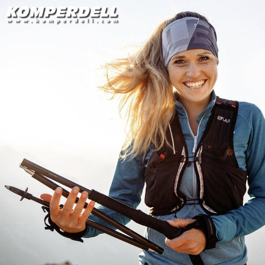A woman in a blue top and charcoal vest holds Komperdell Carbon FXP .4 Approach - Vario Compact trekking poles against a bright outdoor background.