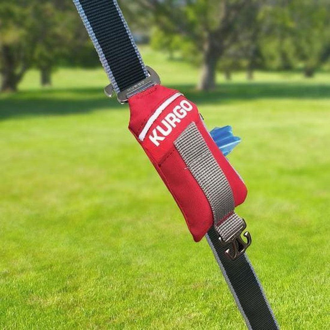 A red Kurgo Duty Bag attached to a black strap with a gray strap, set against a green grassy outdoor background.