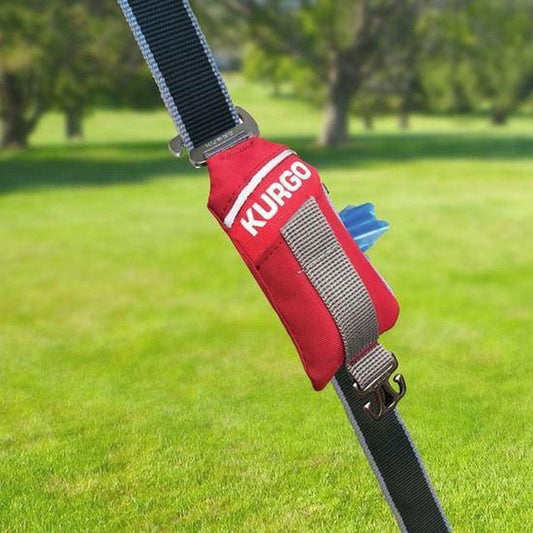 A red Kurgo Duty Bag attached to a black strap with a gray strap, set against a green grassy outdoor background.