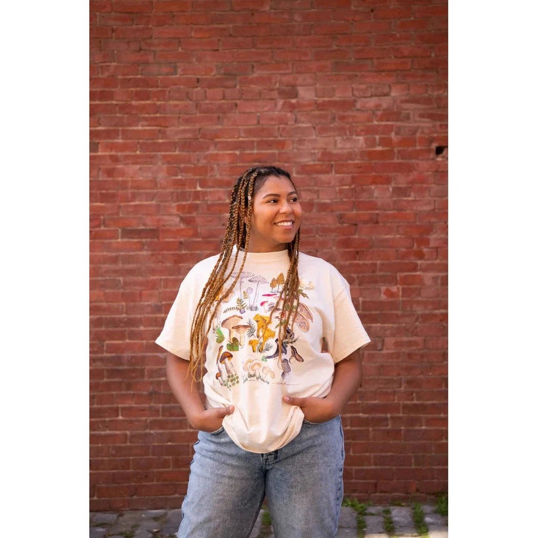 A woman wearing the Natural Liberty Graphics Mushrooms Short Sleeve T stands in front of a red brick wall.