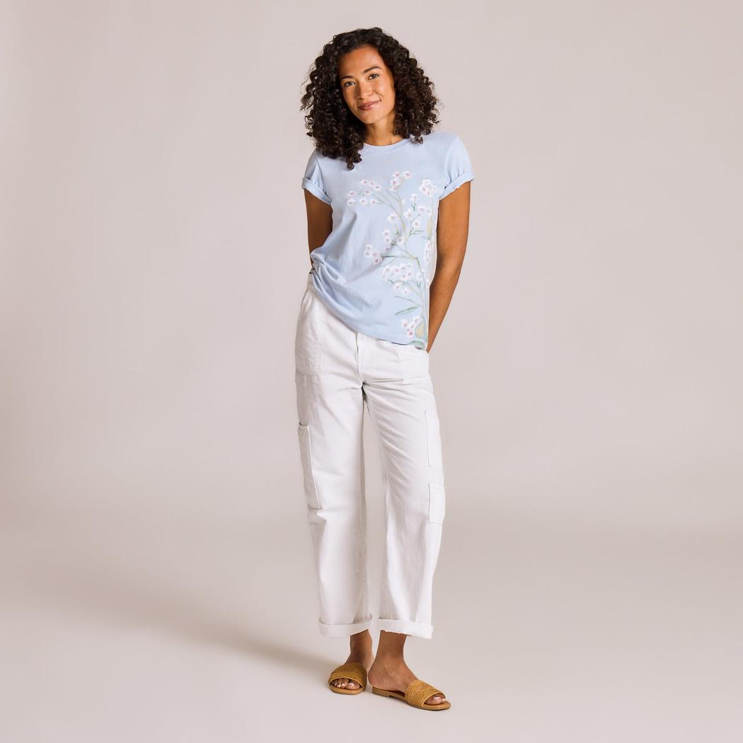 A woman wearing the Life is Good Women's Forgetmenot Butterfly Short Sleeve Crusher Tee in Glacier Blue stands against a plain background.