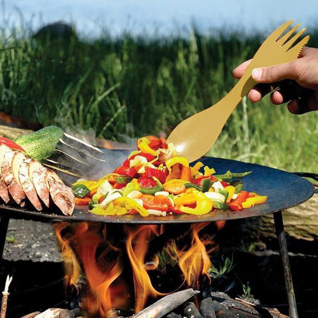 A hand uses the Light My Fire Spork Large Serving BIO to stir colorful veggies on a Hazy Blue camping grill over a campfire with grass in the background.