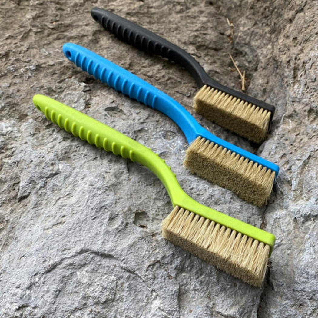 Three Metolius Razorback Boar's Hair Brushes in Blue, Green, and Charcoal/Graphite lie on a rocky surface.