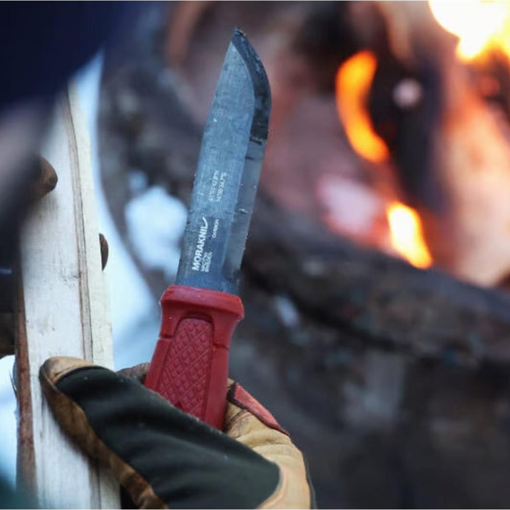 A close-up of the Morakniv Garberg Survival Blackblade knife with a red handle, held in a gloved hand near a fire.