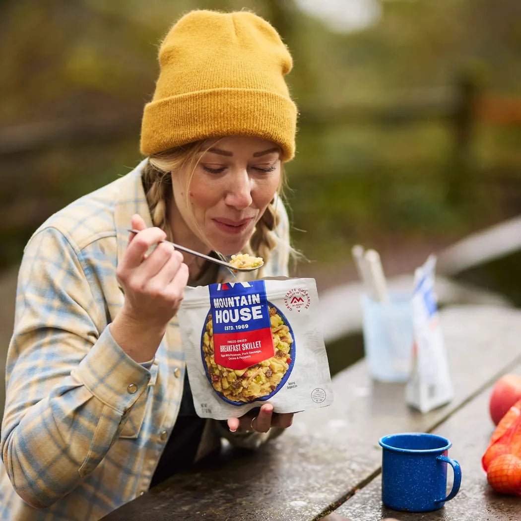 A woman in a yellow beanie eats Mountain House Breakfast Skillet from a blue mug on a wooden table outdoors.