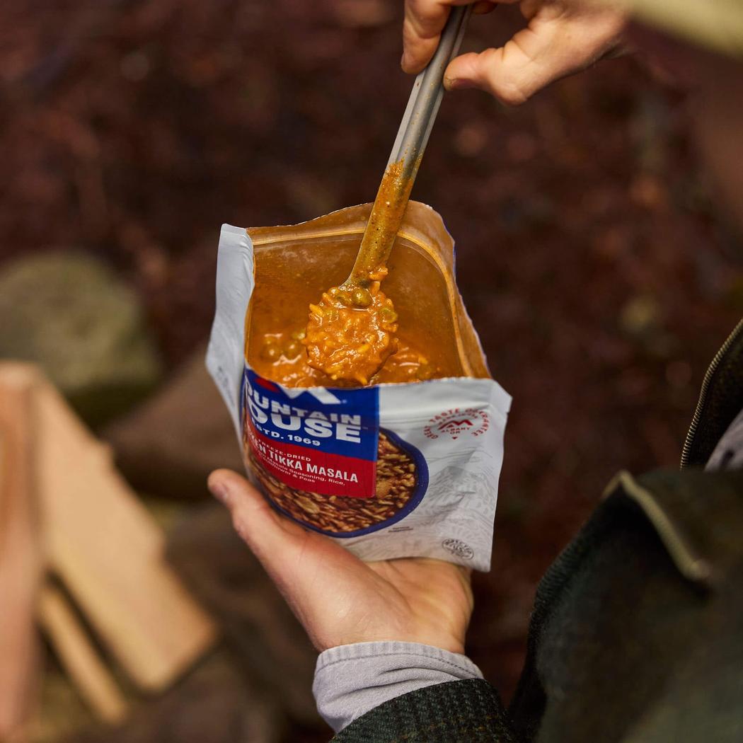 A hand holds a Mountain House Chicken Tikka Masala (GF) pouch while another hand stirs the contents with a spoon, set against a dark, outdoor background.
