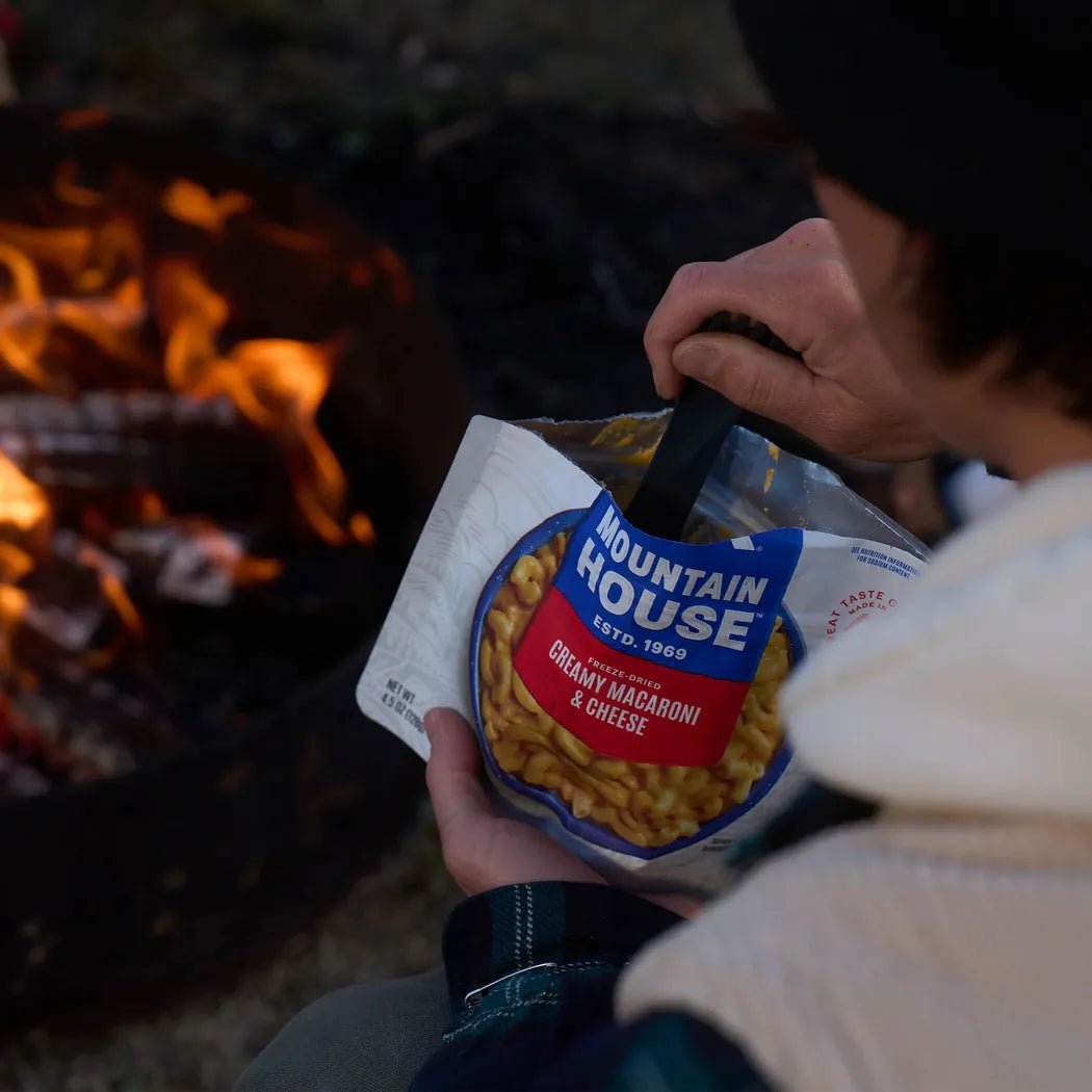 A person holds a Mountain House Creamy Macaroni and Cheese pouch near a campfire.