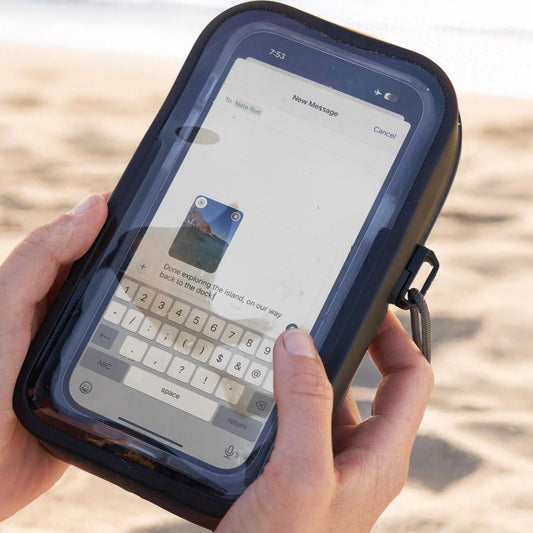 A person holds the black Nite Ize RunOff Waterproof Phone Case Plus on a sandy beach, showing the phone screen and keyboard.