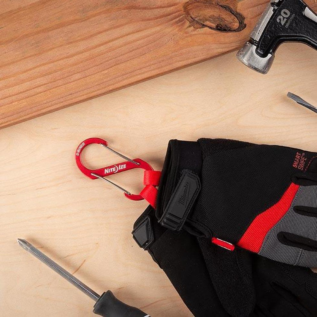 A red Nite Ize S-Biner Aluminum Dual Carabiner #4 hangs from a black and red glove on a light wooden surface with a hammer nearby.