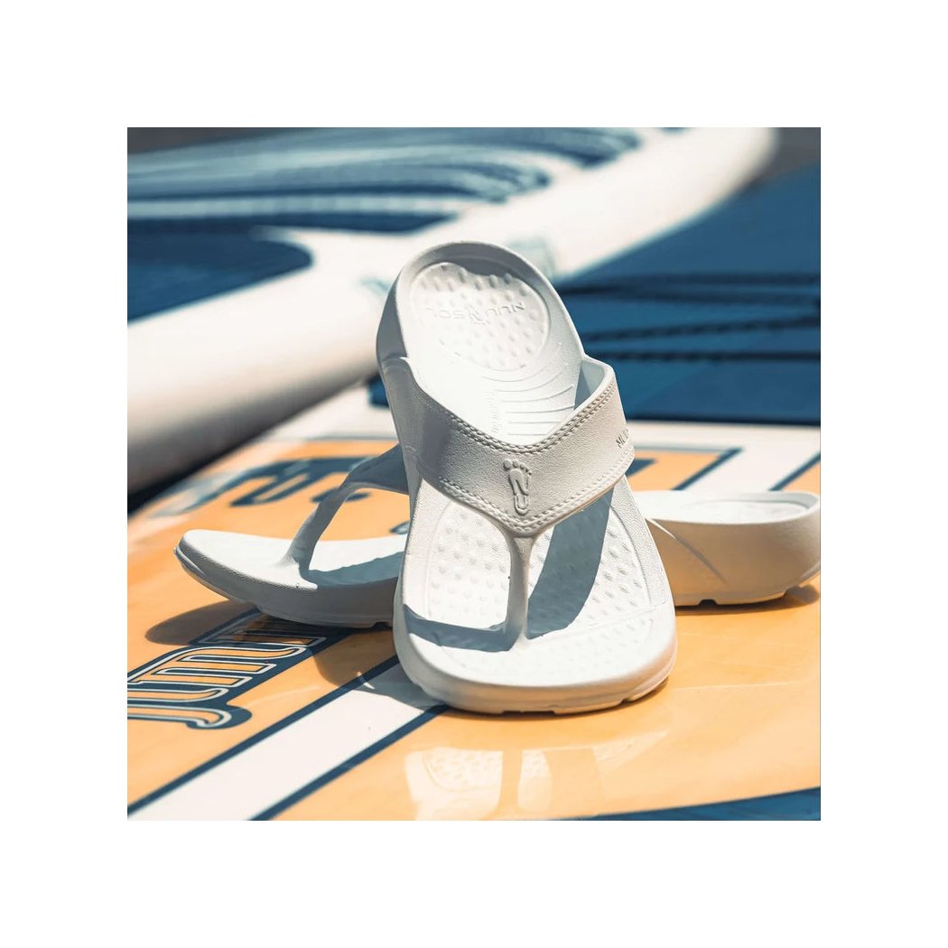 White NuuSol Womens Cascade Flip Flops with a textured footbed sit on a yellow and white surfboard in bright sunlight.