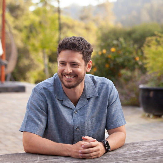 A man wearing the Old Ranch Men's Bodi Shirt in Dark Blue sits at a table outdoors with trees and a potted plant in the background.