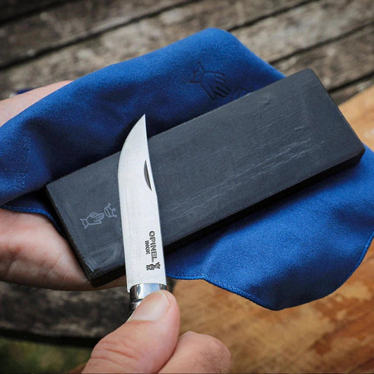 A hand holds a white Opinel knife over a charcoal sharpening stone on a blue cloth, with a wooden background.