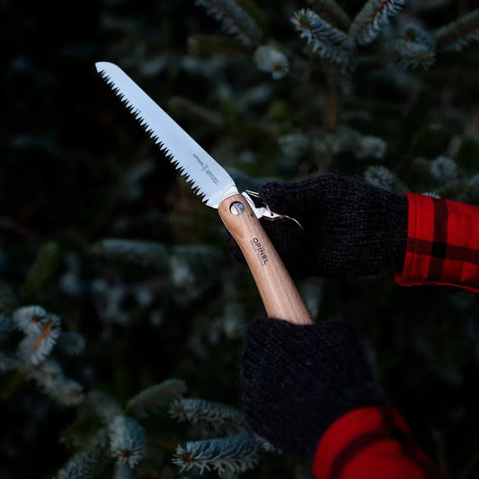 A person wearing black and red plaid gloves holds the Opinel No.18 Carbon Steel Folding Garden Saw in front of a snowy evergreen tree.
