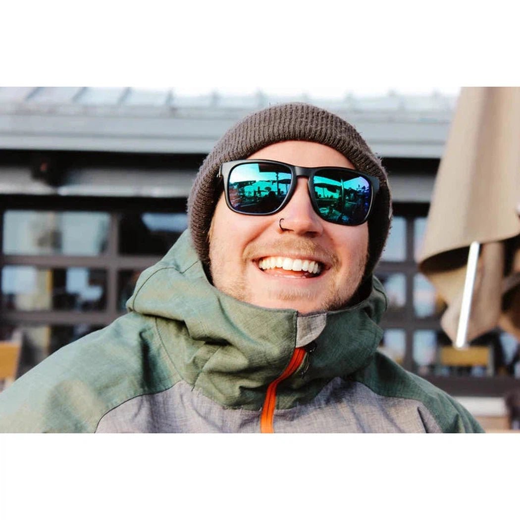 A smiling model wears the Optic Nerve Ziggy sunglasses in Matte Black, with a reflective blue lens, against an outdoor backdrop.