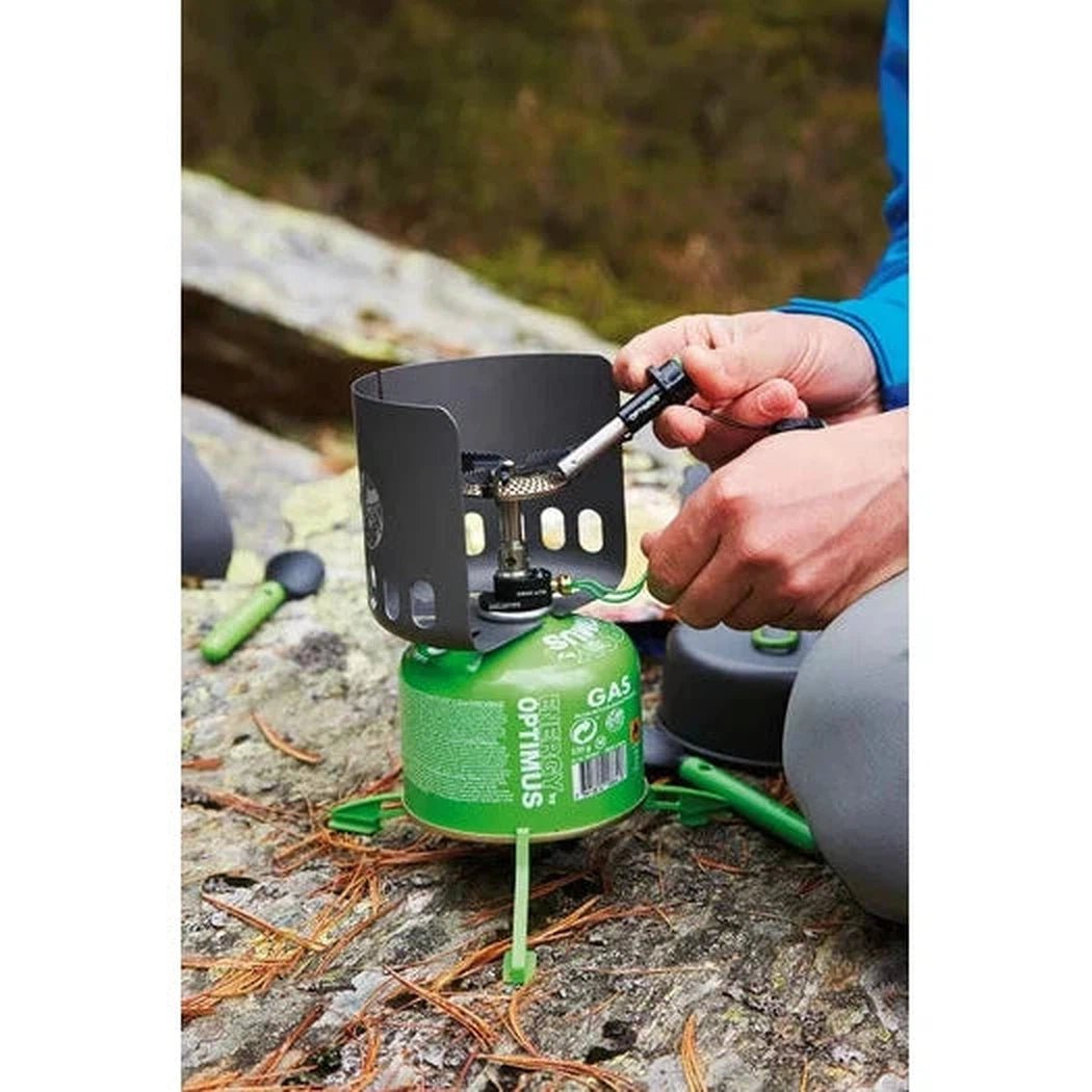 A person lighting the Optimus Sparky camping stove on a rocky outdoor surface with pine needles.