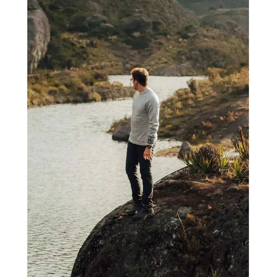 A man stands on a rock by a river wearing the Paka Apparel The Costa in Light Grey, with mountains and water in the background.