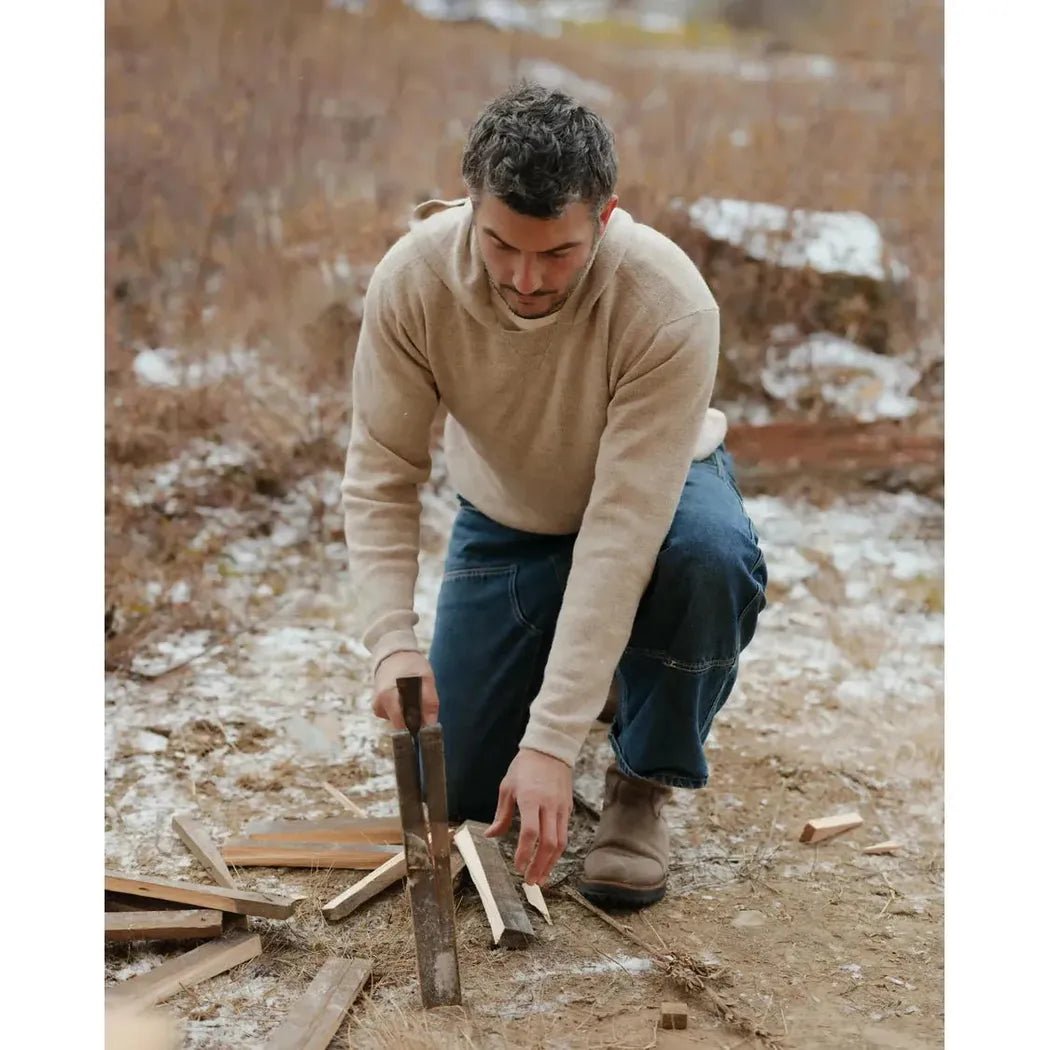 A man in a Pebble Paka Apparel The Hoodie kneels outdoors cutting wood with a saw.