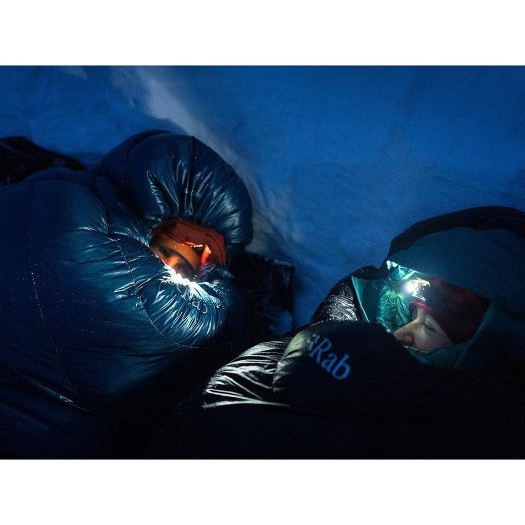 A person wearing a headlamp lies in a Rab Ascent 900 sleeping bag in a snowy, nighttime setting.