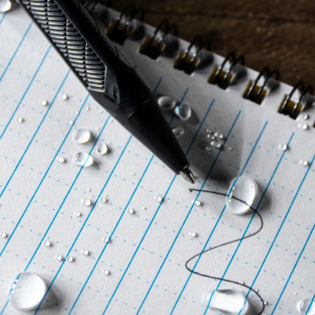 A close-up of the Black Rite in the Rain Mini Bolt-Action Pen on a lined notebook with water droplets.