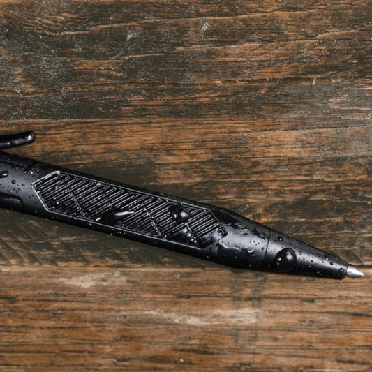 A black Rite in the Rain Mini Bolt-Action Pen lies on a weathered wooden surface with water droplets visible on its body.