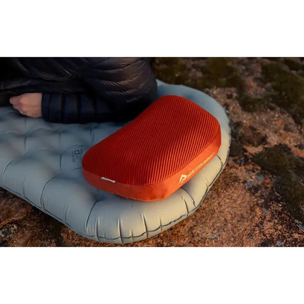 A side view of the Sea To Summit Aeros Premium Pillow in Burnt Olive on a gray sleeping pad with a person’s arm nearby on a rocky surface.