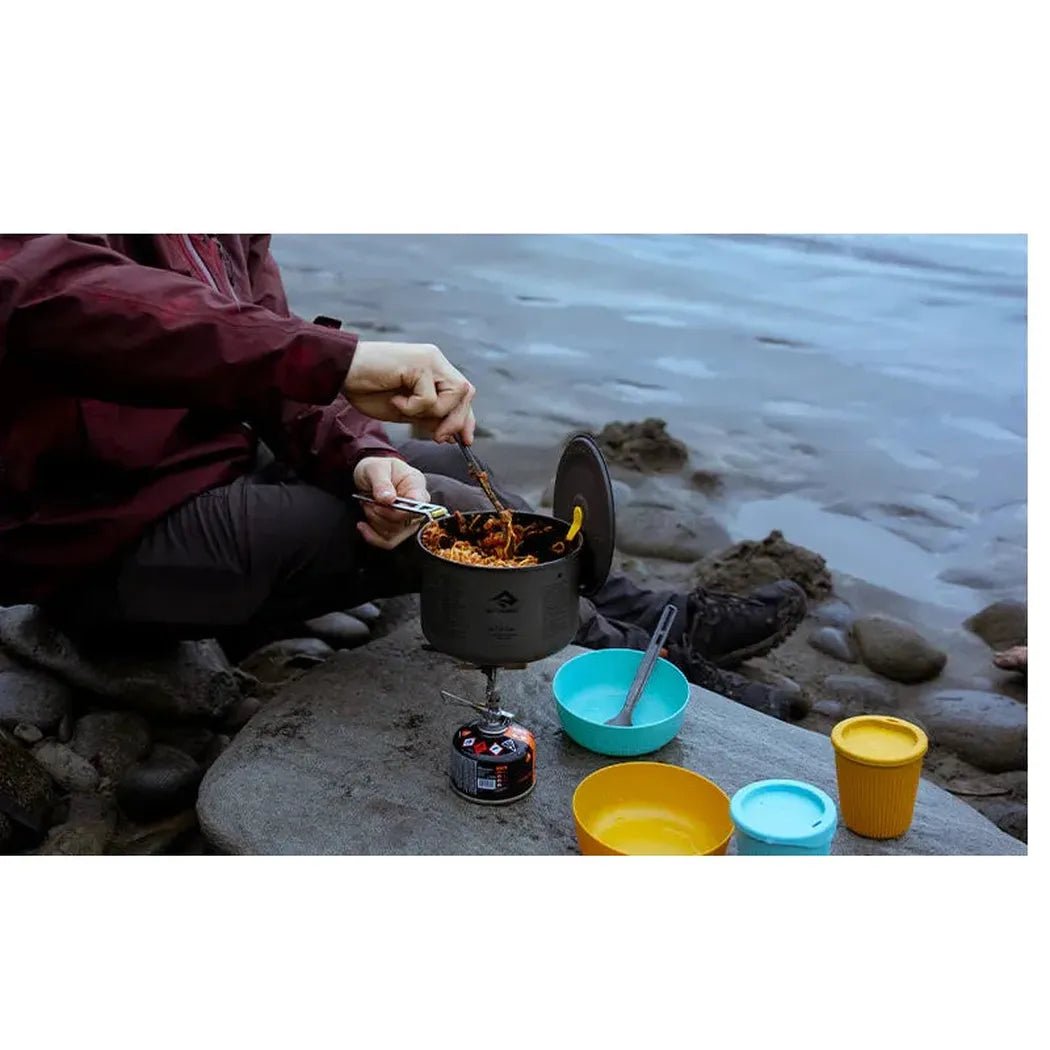 A person cooks on a rocky beach with the Sea To Summit Frontier UL Pot - 1.3L on a portable stove, surrounded by colorful camping dishes.