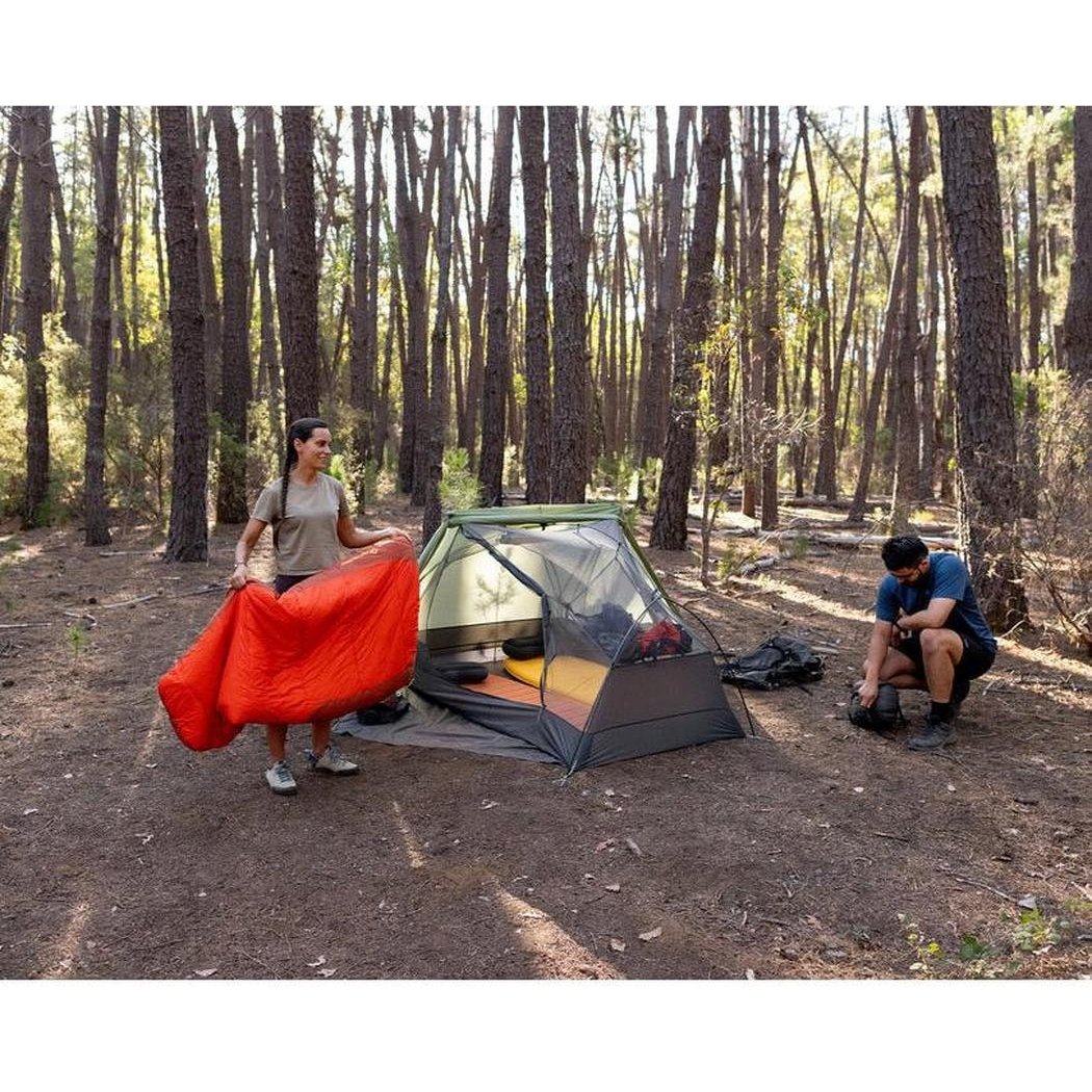 A woman holds the Sea To Summit Hamelin Women's Synthetic Sleeping Bag in a forest with a tent and another person nearby.