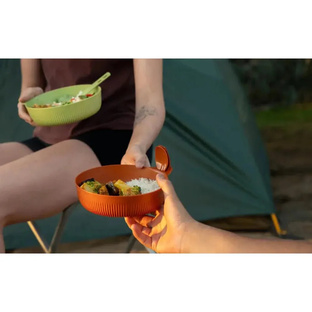 A hand holds a red Sea To Summit Horizon Bowl - L with food inside, while another person sits by a green tent with a matching bowl.