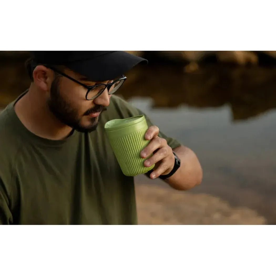 A man in a black cap and olive shirt holds the Sea To Summit Horizon Insulated Mug - 475ml in Bombay color, drinking from it near a body of water.