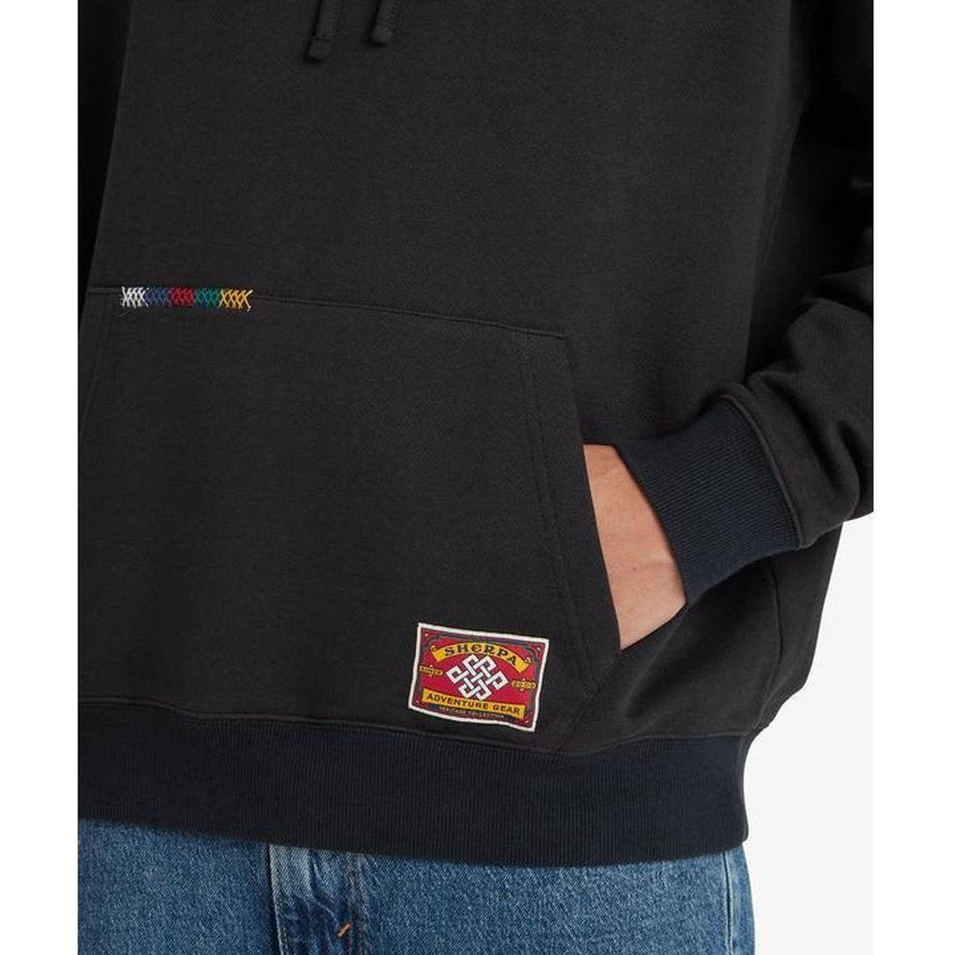 A close-up of the Sherpa Adventure Gear Men's Tarcho Mask Hoodie in Black, showing the front pocket and a colorful patch on the chest.