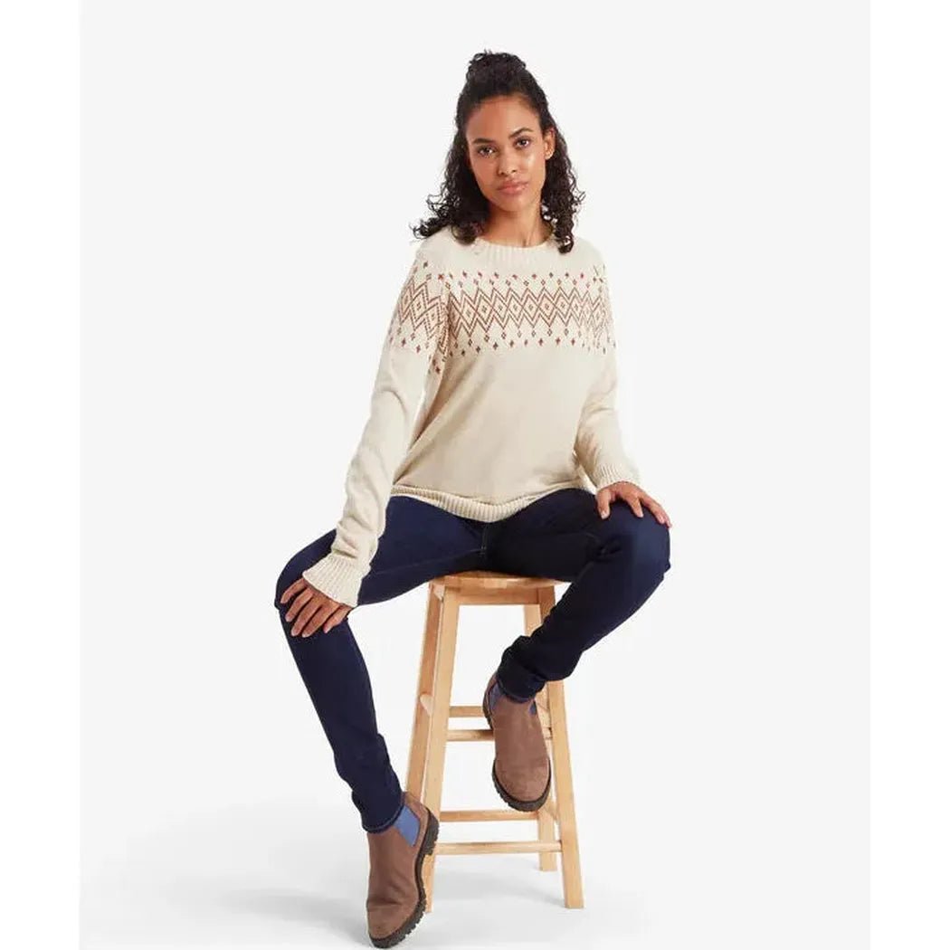 A woman sits on a wooden stool wearing the Sherpa Adventure Gear Samples - Women's Yeshe Crew Neck Sweater in Ecru against a white background.
