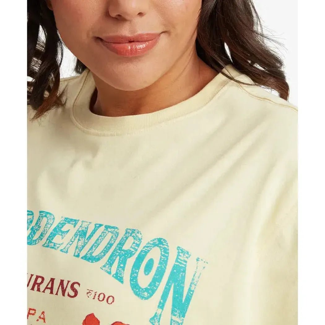 A close-up of a woman wearing the Sherpa Adventure Gear Women's Tarcho Rhodoendron Tee in Butter color, showing the front graphic design against a white background.