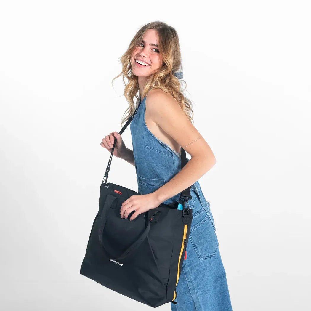 A woman in blue overalls carries the Sherpani Jetsetter Tote in Chromatic against a white background.