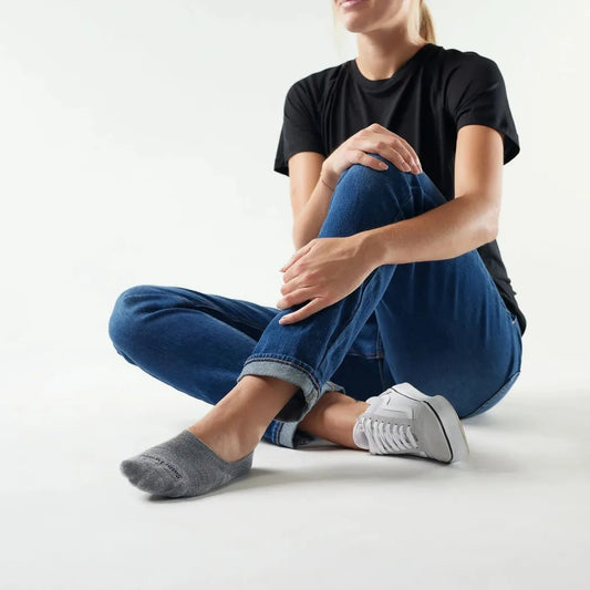 A woman sits cross-legged wearing Smartwool Everyday No Show Socks in Medium Gray with a white sneaker and blue jeans against a white background.