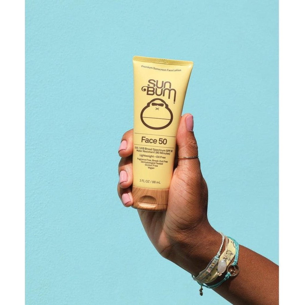 A hand holds a yellow Sun Bum Face 50 sunscreen tube against a light blue background.