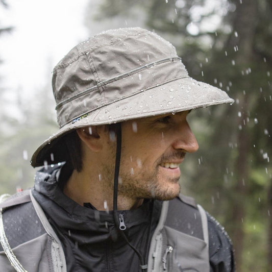 A man wears the Sunday Afternoons Ultra Storm Bucket hat in Carbon color, with raindrops visible on the brim and background showing trees.