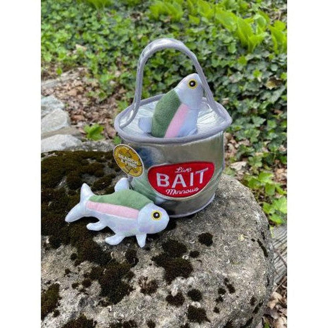 A Tall Tails Bait Bucket Squeaky Hide & Seek Toy sits on a mossy rock with a matching fish toy beside it, surrounded by green foliage.