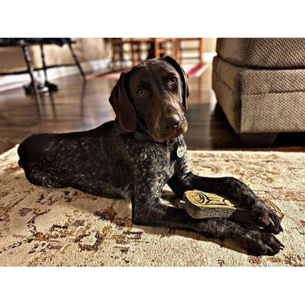 A charcoal dog lies on a rug with its Tall Tails Natural Leather & Wool Love My Dog Log toy.
