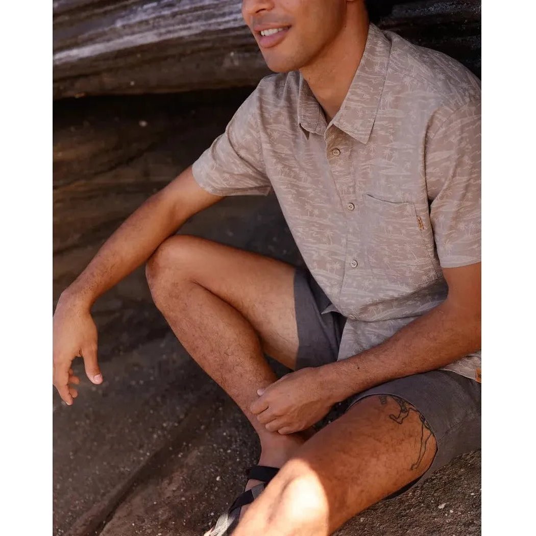 A man wearing the Tentree Men's Redwood Pocket Shirt in Khaki/Palm sits outdoors on rocks with a log behind him.