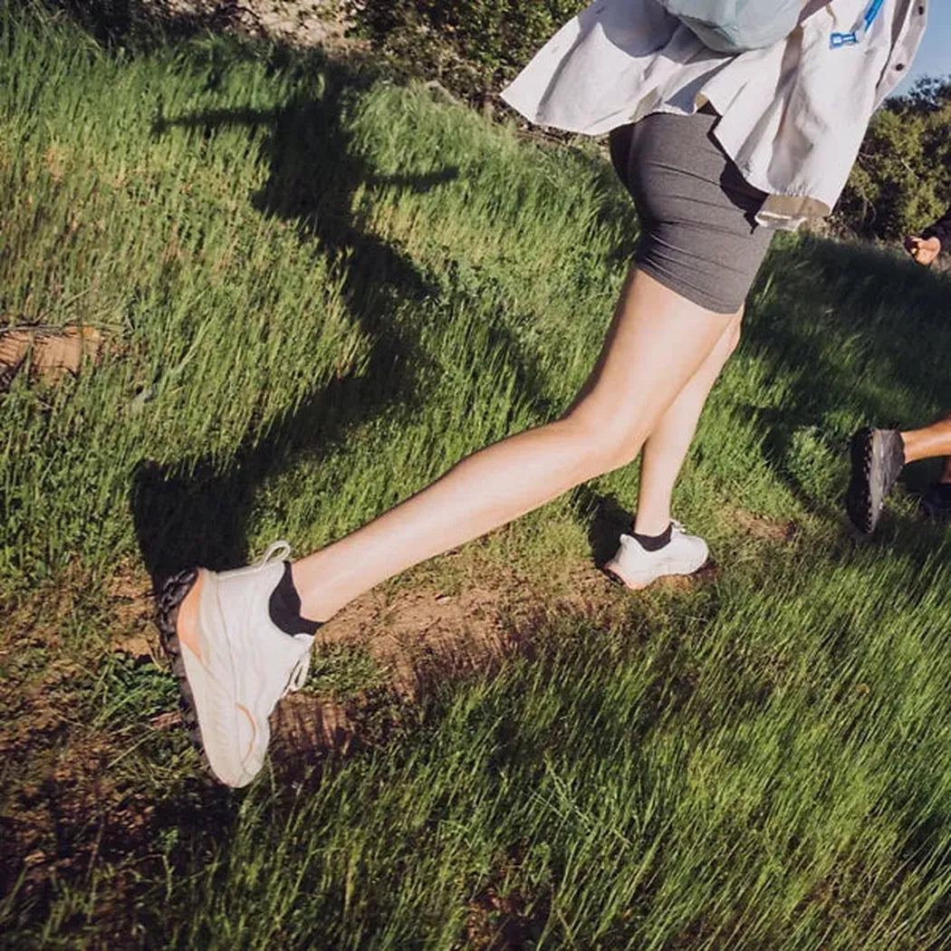 A woman wearing the Teva Women's Trailwinder Low in Birch/ Peach Bloom runs on a grassy trail.
