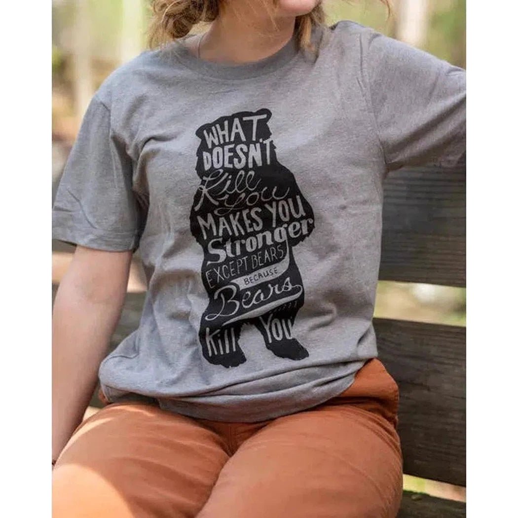 A woman wearing a Smoke Grey The Landmark Project Bear Tee sits on a wooden bench outdoors.