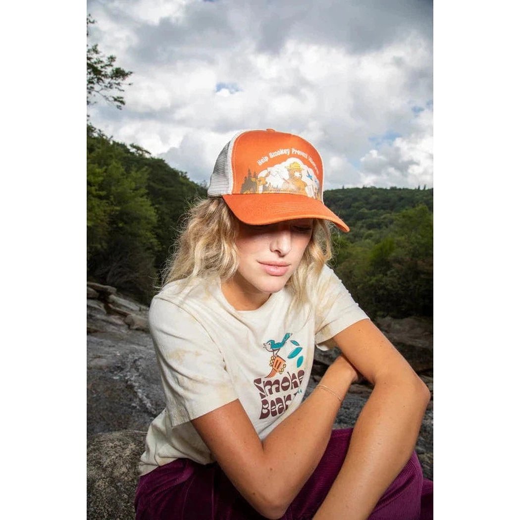 A woman wearing The Landmark Project Help Smokey 5-Panel Trucker Hat in Terracotta sits on rocks with a forest and cloudy sky behind her.