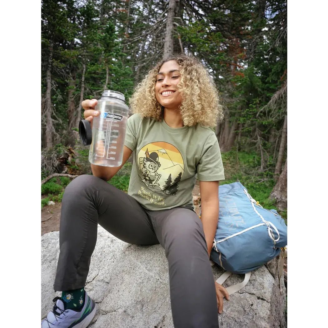 A woman in an olive The Landmark Project Pack It in, Pack It Out tee sits on a rock in a forest, holding a water bottle.