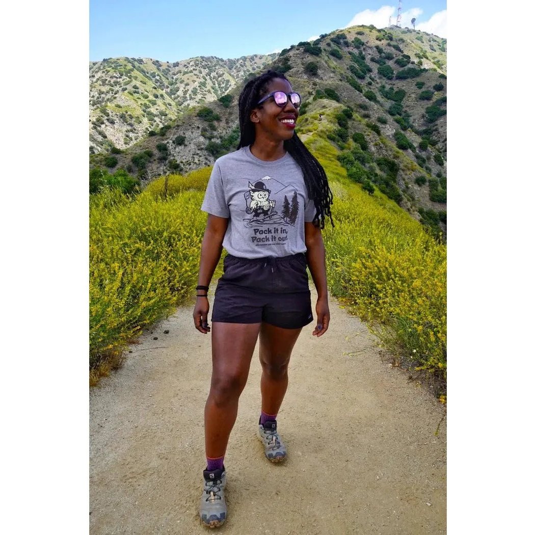 A woman wearing The Landmark Project Pack It in, Pack It Out smoke grey tee and black shorts smiles on a hiking trail with green hills in the background.