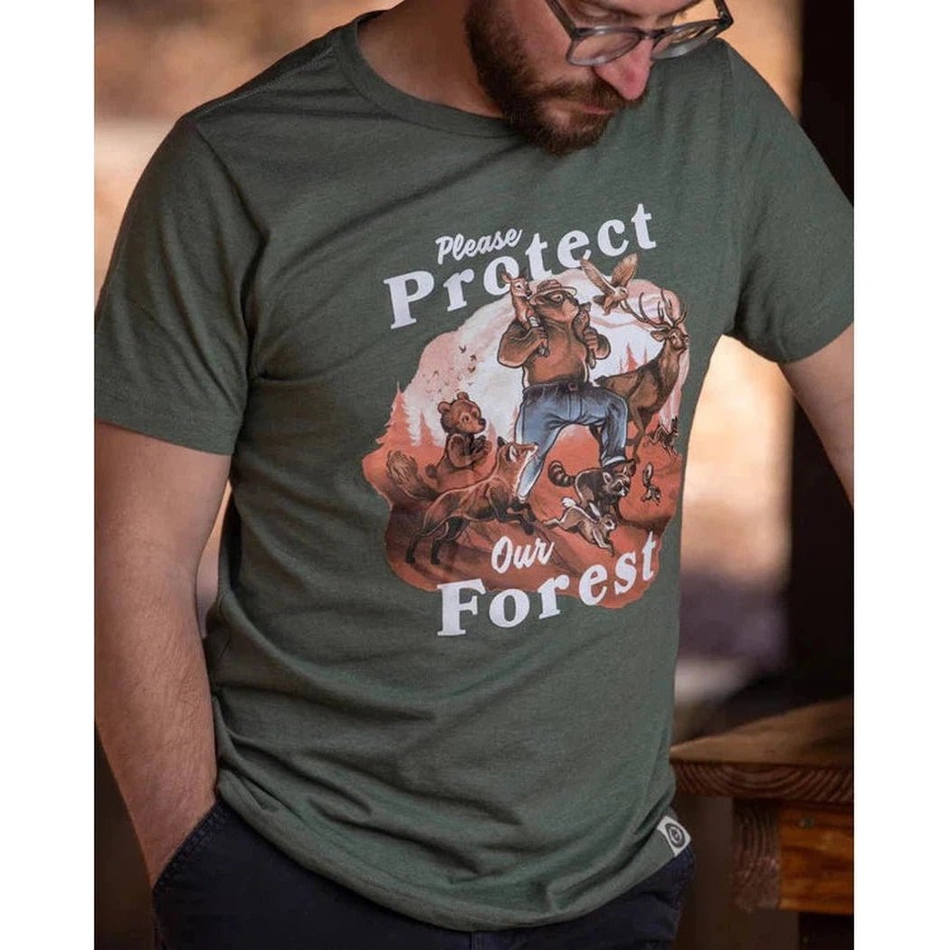 A man wearing a Conifer The Landmark Project Protect Our Forest Tee looks down at a wooden table in the background.