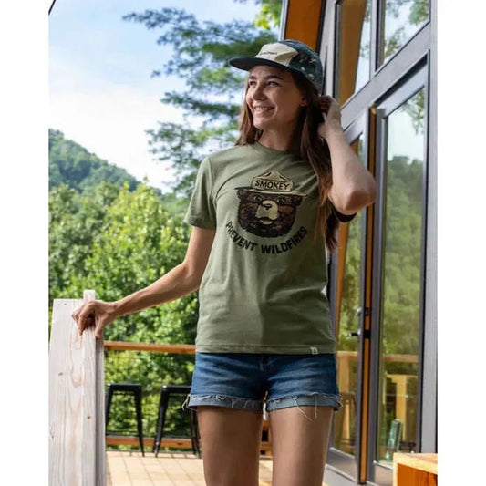 A woman wearing The Landmark Project Smokey Retro Unisex Short Sleeve Tee in Cactus stands on a deck with trees in the background.