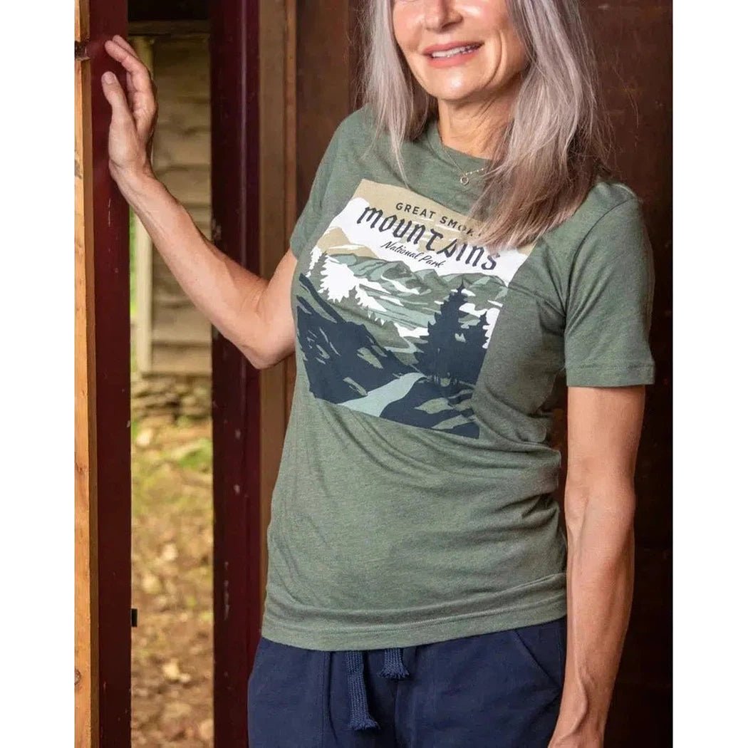 A woman wearing The Landmark Project Smoky Mountains National Park Tee in Conifer stands by a door with a mountain graphic on the tee.